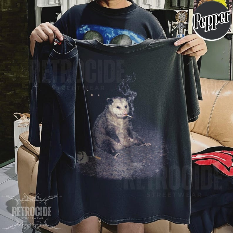 K&ouml;nnte beinhalten: Dunkelgraues T-Shirt mit einer Grafik eines Opossums, das eine Zigarette raucht. Das Opossum sitzt, und Rauch steigt von der Zigarette auf. Das Shirt tr&auml;gt den Text "RETROCIDE STREETWEAR".