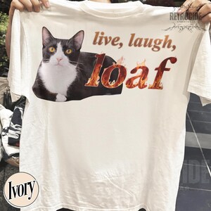 Puede incluir: Una camiseta de color marfil con un gr&aacute;fico de un gato blanco y negro tumbado. El texto "live, laugh, loaf" est&aacute; impreso encima y al lado del gato, con la palabra "loaf" representada en un patr&oacute;n de llamas de color naranja y rojo. El nombre de la marca "RETROCIDE" es visible en la esquina superior derecha.