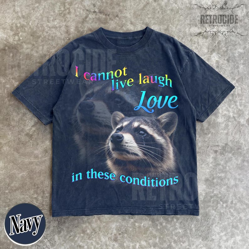 Puede incluir: Una camiseta de manga corta en color azul marino oscuro con un gr&aacute;fico de dos mapaches. El texto dice I cannot live laugh Love in these conditions en un degradado de colores azul, morado y amarillo. La camiseta tiene cuello redondo y corte holgado.