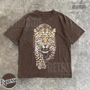 May include: Espresso-colored t-shirt featuring a detailed leopard graphic. The leopard is depicted in shades of brown and tan, with a fierce expression. The shirt has a classic crew neck and short sleeves. The brand name "RETROCIDE STREETWEAR" is printed on the shirt.