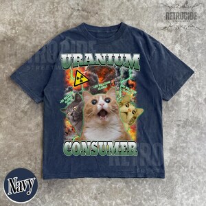 Puede incluir: Una camiseta azul marino con un estampado gr&aacute;fico de tres gatos sobre un fondo de fuego y rayos verdes. El texto dice URANIUM CONSUMER y 20 BILLION CLORIC. Un s&iacute;mbolo amarillo de advertencia de radiaci&oacute;n se sit&uacute;a sobre los gatos.