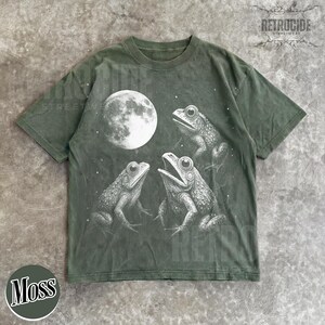 May include: Moss green t-shirt featuring a graphic of three frogs under a full moon. The frogs are detailed in white, with the moon also in white. The shirt has a crew neck and short sleeves. The word "Moss" is printed on a green circle.