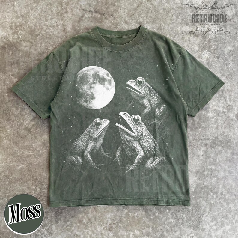 May include: Moss green t-shirt featuring a graphic of three frogs under a full moon. The frogs are detailed in white, with the moon also in white. The shirt has a crew neck and short sleeves. The word "Moss" is printed on a green circle.