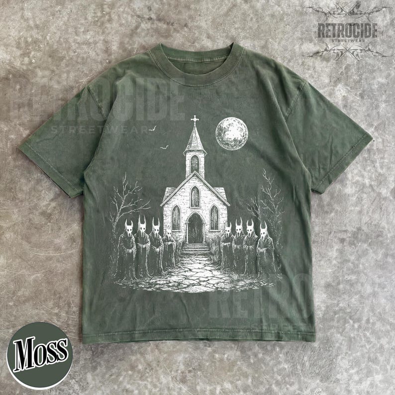 Op de afbeelding: Een mosgroen t-shirt met korte mouwen en een witte grafische print van een kerk met een toren, een volle maan en een groep figuren in capuchons met gehoornde maskers op een stenen pad voor het gebouw, omringd door kale bomen.