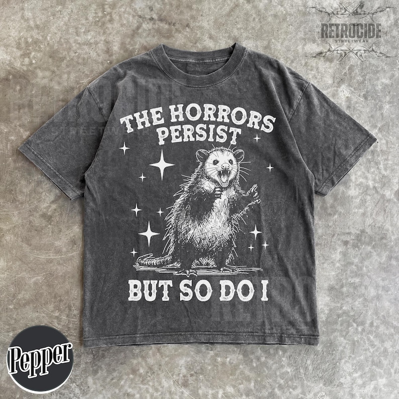 May include: Dark grey t-shirt with a distressed graphic of an opossum and the text "THE HORRORS PERSIST BUT SO DO I". The design includes stars and is from the brand "RETROCIDE STREETWEAR".