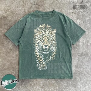 May include: Light green t-shirt featuring a detailed leopard graphic. The leopard is depicted in shades of brown, black, and white, with a fierce expression. The shirt has a classic crew neck and short sleeves. The text "RETROCIDE" is printed on the top right.