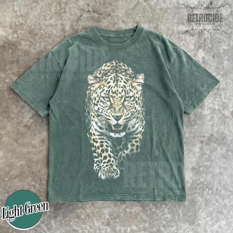 May include: Light green t-shirt featuring a detailed leopard graphic. The leopard is depicted in shades of brown, black, and white, with a fierce expression. The shirt has a classic crew neck and short sleeves. The text "RETROCIDE" is printed on the top right.