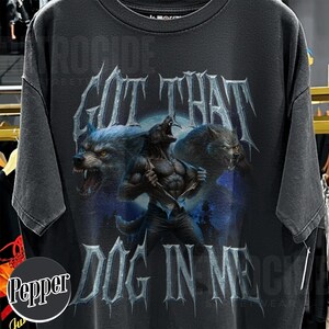 Peut inclure: T-shirt gris fonc&eacute; avec un motif de loup-garou et le texte "GOT THAT DOG IN ME". Le design pr&eacute;sente deux loups hurlants et un loup-garou en transformation sur fond de clair de lune bleu.