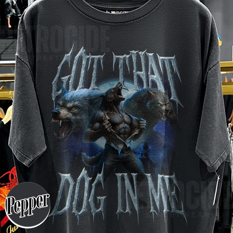 Peut inclure: T-shirt gris fonc&eacute; avec un motif de loup-garou et le texte "GOT THAT DOG IN ME". Le design pr&eacute;sente deux loups hurlants et un loup-garou en transformation sur fond de clair de lune bleu.