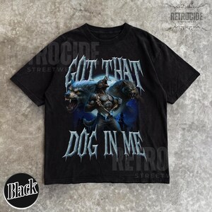 Peut inclure: T-shirt noir avec un motif de loup-garou se transformant sous la pleine lune et le texte "GOT THAT DOG IN ME". Le design a un style streetwear vintage. Col rond classique.