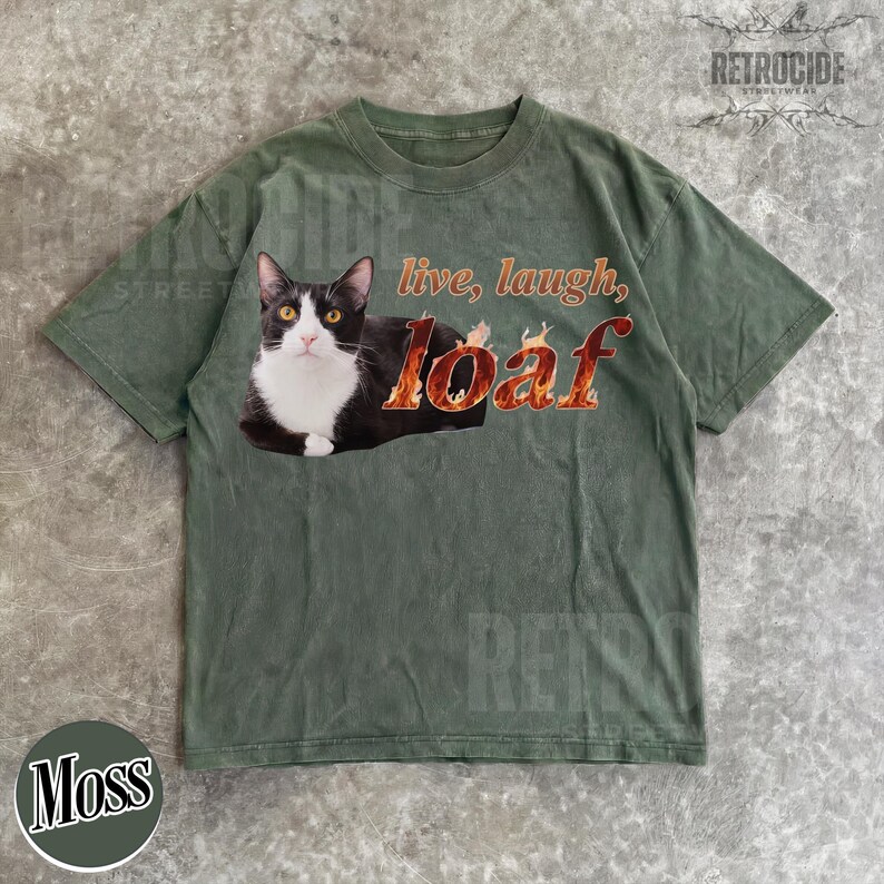 Puede incluir: Una camiseta de manga corta color verde oliva con el gr&aacute;fico de un gato blanco y negro. El texto "live, laugh, loaf" est&aacute; impreso en una fuente estilizada con un patr&oacute;n de llamas. El gato est&aacute; situado a la izquierda del texto en el pecho.