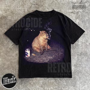 May include: A black short-sleeve t-shirt featuring a graphic print of a capybara sitting on the ground with a lit cigarette in its mouth and a can of Pabst Blue Ribbon beer nearby. The design includes wisps of smoke rising from the cigarette.
