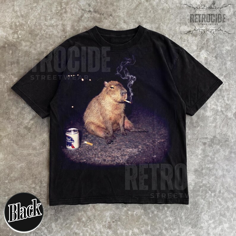 May include: A black short-sleeve t-shirt featuring a graphic print of a capybara sitting on the ground with a lit cigarette in its mouth and a can of Pabst Blue Ribbon beer nearby. The design includes wisps of smoke rising from the cigarette.