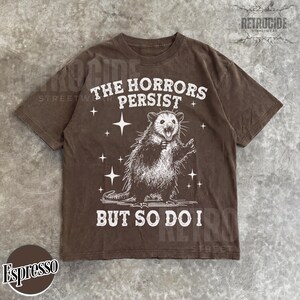 May include: Espresso-colored t-shirt featuring a distressed graphic of an opossum with the text "THE HORRORS PERSIST BUT SO DO I." The design includes stars. The shirt has a crew neck and short sleeves.