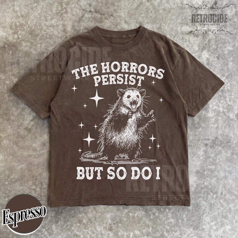 May include: Espresso-colored t-shirt featuring a distressed graphic of an opossum with the text "THE HORRORS PERSIST BUT SO DO I." The design includes stars. The shirt has a crew neck and short sleeves.