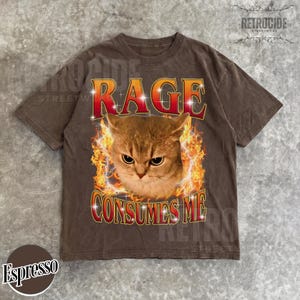 Puede incluir: Una camiseta marr&oacute;n de manga corta con un gr&aacute;fico de un gato rodeado de llamas naranjas y amarillas. El texto RAGE est&aacute; impreso en rojo sobre el gato, y CONSUMES ME est&aacute; impreso en rojo debajo del gato, con peque&ntilde;os detalles de estrellas.