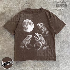 May include: Espresso-colored t-shirt featuring a detailed illustration of three frogs and a full moon. The graphic is in white, creating a striking contrast against the dark brown fabric. The shirt has a classic crew neck and short sleeves.