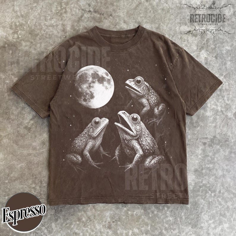 May include: Espresso-colored t-shirt featuring a detailed illustration of three frogs and a full moon. The graphic is in white, creating a striking contrast against the dark brown fabric. The shirt has a classic crew neck and short sleeves.