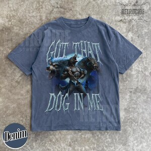 Peut inclure: T-shirt bleu denim avec un motif de loup-garou et le texte "GOT THAT DOG IN ME". Le t-shirt a un col rond et des manches courtes. La marque "RETROCIDE" est imprim&eacute;e en haut.