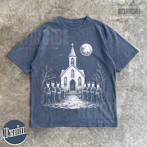 Op de afbeelding: Een leiblauw t-shirt met korte mouwen en een witte grafische print van een kerk onder een volle maan met een groep figuren in capuchons ervoor. Het ontwerp bevat kale bomen en een stenen pad dat naar de ingang van de kerk leidt.