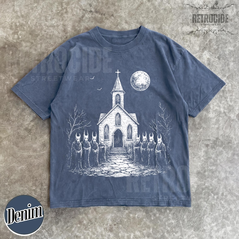 Op de afbeelding: Een leiblauw t-shirt met korte mouwen en een witte grafische print van een kerk onder een volle maan met een groep figuren in capuchons ervoor. Het ontwerp bevat kale bomen en een stenen pad dat naar de ingang van de kerk leidt.