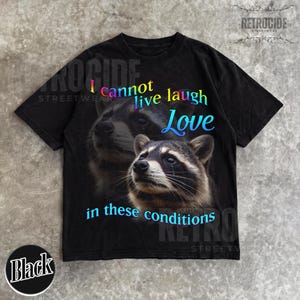 Puede incluir: Una camiseta negra de manga corta con un gr&aacute;fico de dos mapaches. El texto "I cannot live laugh Love in these conditions" est&aacute; impreso en un degradado de azul, morado y amarillo. El dise&ntilde;o incluye una cara de mapache grande y detallada.