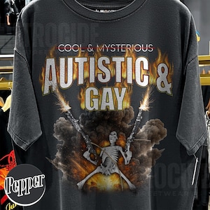 May include: Dark gray t-shirt with the text "COOL & MYSTERIOUS AUTISTIC & GAY" in a distressed, fiery font. A skeleton figure is depicted holding two guns with explosions in the background. The shirt has a vintage, worn look.