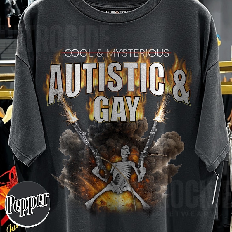 May include: Dark gray t-shirt with the text "COOL & MYSTERIOUS AUTISTIC & GAY" in a distressed, fiery font. A skeleton figure is depicted holding two guns with explosions in the background. The shirt has a vintage, worn look.