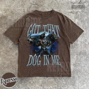 Peut inclure: T-shirt couleur expresso avec un motif de loup-garou et l'inscription "GOT THAT DOG IN ME". Le motif comprend deux loups hurlants. Col rond, manches courtes.