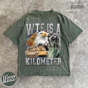 May include: Moss green t-shirt with a graphic design featuring a hot dog, eagle, raccoon, tank, and the Statue of Liberty. The text "WTF IS A KILOMETER" is printed in white across the front. The shirt has short sleeves and a relaxed fit.