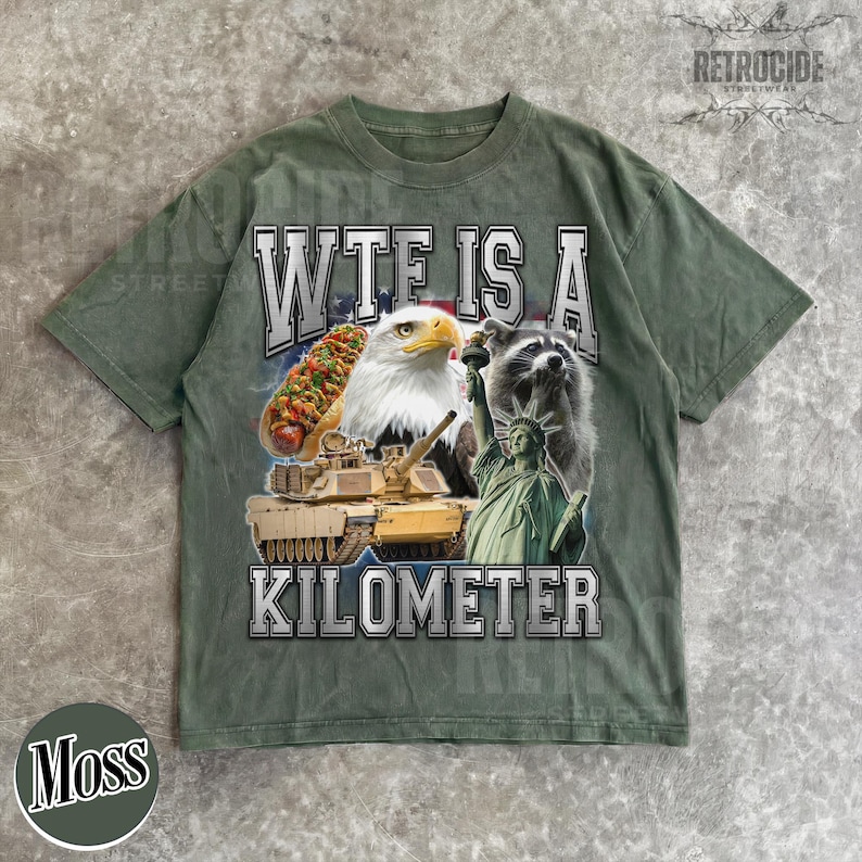May include: Moss green t-shirt with a graphic design featuring a hot dog, eagle, raccoon, tank, and the Statue of Liberty. The text "WTF IS A KILOMETER" is printed in white across the front. The shirt has short sleeves and a relaxed fit.