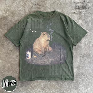 May include: A dark olive green short-sleeve t-shirt featuring a graphic print of a capybara sitting on the ground with a lit cigarette in its mouth, smoke rising above, and a silver beer can with a blue and red label placed to its left side.