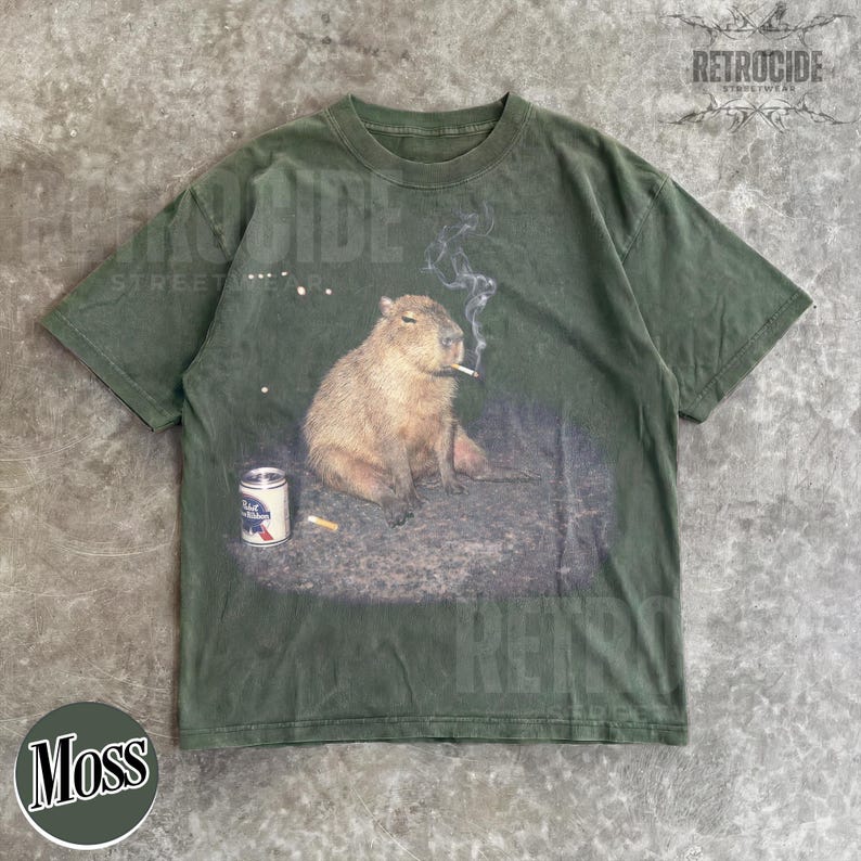 May include: A dark olive green short-sleeve t-shirt featuring a graphic print of a capybara sitting on the ground with a lit cigarette in its mouth, smoke rising above, and a silver beer can with a blue and red label placed to its left side.