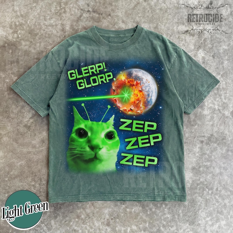 May include: Light green t-shirt with a graphic of a green cat with antennae, a planet being zapped by a laser, and the words "GLERP! GLORP." and "ZEP ZEP ZEP." The shirt has a vintage, washed look.