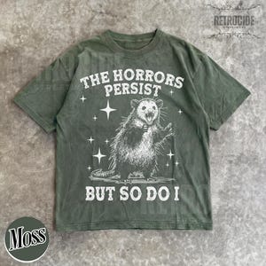 May include: Moss green t-shirt with a distressed graphic of an opossum and the text "THE HORRORS PERSIST BUT SO DO I". The design includes stars around the opossum. The shirt has a crew neck and short sleeves.