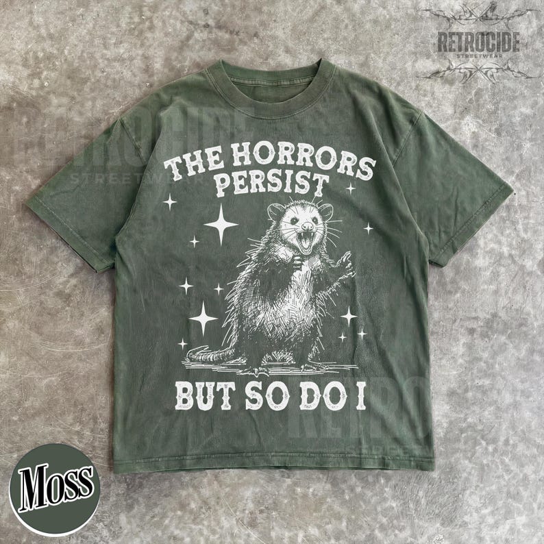 May include: Moss green t-shirt with a distressed graphic of an opossum and the text "THE HORRORS PERSIST BUT SO DO I". The design includes stars around the opossum. The shirt has a crew neck and short sleeves.