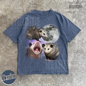 May include: Denim blue t-shirt with a graphic of three opossums, a full moon, and a galaxy background. The shirt has a crew neck and short sleeves. The word "Denim" is printed on the bottom left.