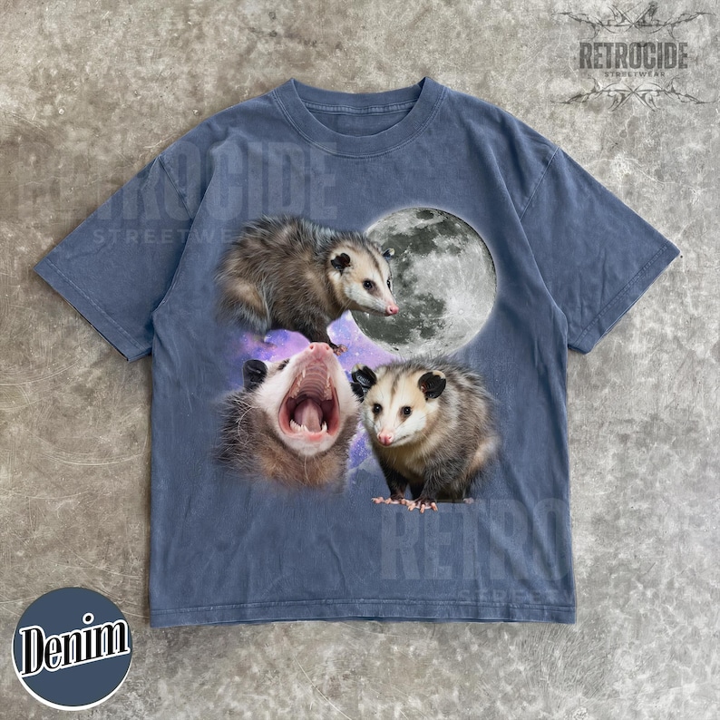May include: Denim blue t-shirt with a graphic of three opossums, a full moon, and a galaxy background. The shirt has a crew neck and short sleeves. The word "Denim" is printed on the bottom left.