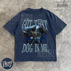 Peut inclure: T-shirt bleu marine avec un motif de loup-garou et le texte "GOT THAT DOG IN ME". Le design comprend un loup-garou et deux loups mena&ccedil;ants. Le t-shirt a un aspect vintage et vieilli.