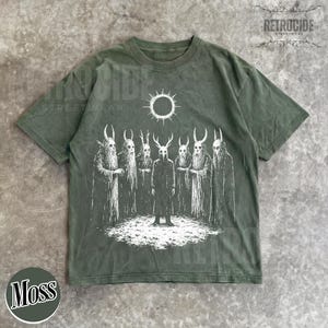 May include: Moss green t-shirt with a white graphic of figures with horned masks and a sun. The shirt has a crew neck and short sleeves. The word "RETROCIDE" is printed on the top right corner.