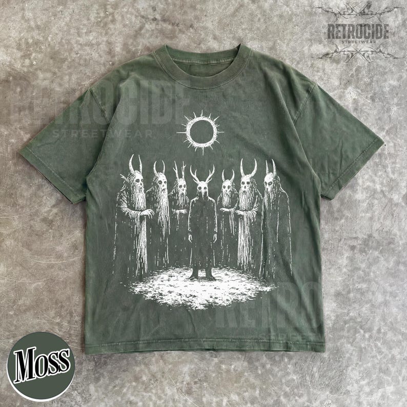May include: Moss green t-shirt with a white graphic of figures with horned masks and a sun. The shirt has a crew neck and short sleeves. The word "RETROCIDE" is printed on the top right corner.