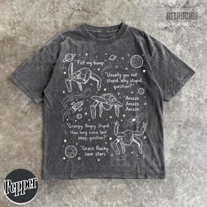 May include: A charcoal gray short-sleeve t-shirt featuring white line art of space creatures, planets, and stars. The graphic includes text: Fist my bump, Usually you not stupid. Why stupid, question?, Amaze. Amaze. Amaze., Grumpy. Angry. Stupid. How long since last sleep, question?, Grace Rocky save stars.
