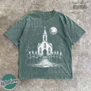 Op de afbeelding: Een lichtgroen t-shirt met korte mouwen en een witte grafische print van een kerk met een toren, een volle maan en een groep gehoornde figuren in mantels voor het gebouw, met kale bomen en een stenen pad in het ontwerp.
