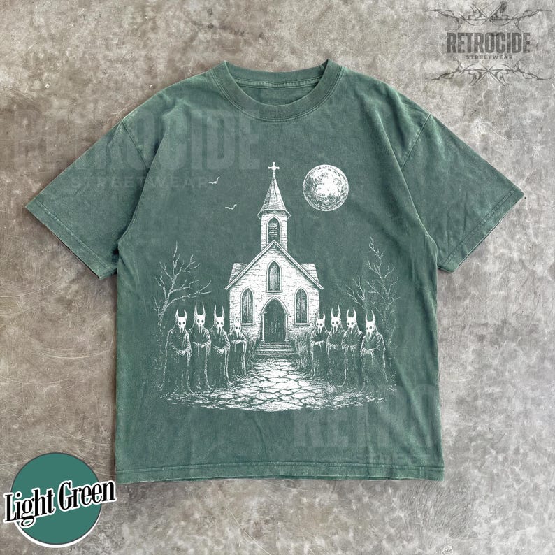 Op de afbeelding: Een lichtgroen t-shirt met korte mouwen en een witte grafische print van een kerk met een toren, een volle maan en een groep gehoornde figuren in mantels voor het gebouw, met kale bomen en een stenen pad in het ontwerp.