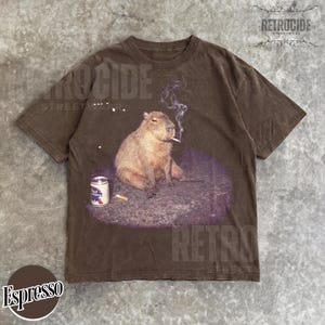 May include: A brown short-sleeve t-shirt featuring a graphic of a capybara sitting on a dark patch, smoking a cigarette with visible smoke, and a Pabst Blue Ribbon beer can next to it. The shirt has a crew neckline and a relaxed fit design.