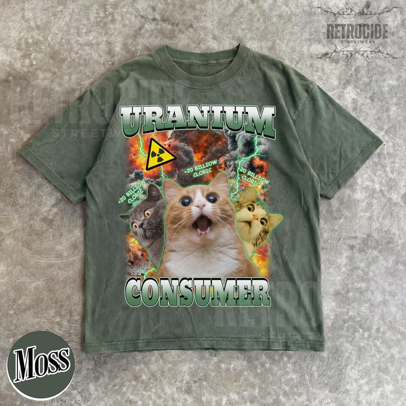 Puede incluir: Una camiseta verde musgo con un estampado gr&aacute;fico de tres gatos sobre un fondo de explosi&oacute;n. El texto dice "URANIUM CONSUMER" con "20 BILLION CLORIC" repetido. Un s&iacute;mbolo amarillo de advertencia de radiaci&oacute;n se ve sobre el gato central.
