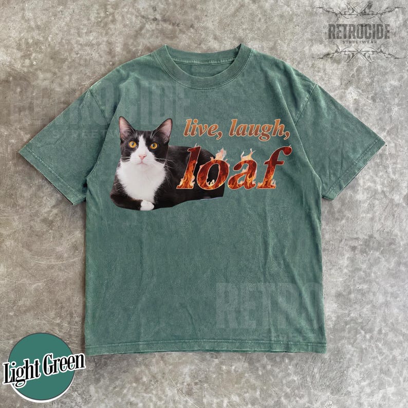 Puede incluir: Una camiseta de manga corta color verde salvia con el gr&aacute;fico de un gato blanco y negro tumbado. El texto "live, laugh, loaf" est&aacute; impreso en un patr&oacute;n de llamas naranja y amarillo junto al gato. La camiseta tiene un cuello redondo.