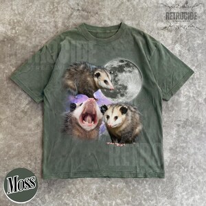 May include: Moss green t-shirt with a graphic of three opossums, a full moon, and a galaxy background. One opossum is yawning. The shirt has a vintage, distressed appearance. The word "Moss" is printed on the bottom left.