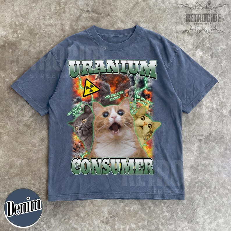 Puede incluir: Una camiseta de manga corta en azul deste&ntilde;ido con un estampado gr&aacute;fico de tres gatos sobre un fondo de fuego y rayos. El texto dice URANIUM CONSUMER con un s&iacute;mbolo de radiaci&oacute;n amarillo y la frase 20 BILLION CLORIC en color verde.