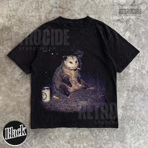 K&ouml;nnte beinhalten: Schwarzes T-Shirt mit einem Aufdruck eines Opossums, das eine Zigarette raucht, mit einer Dose und einer Zigarettenkippe. Auf dem Shirt steht "RETROCIDE STREETWEAR".